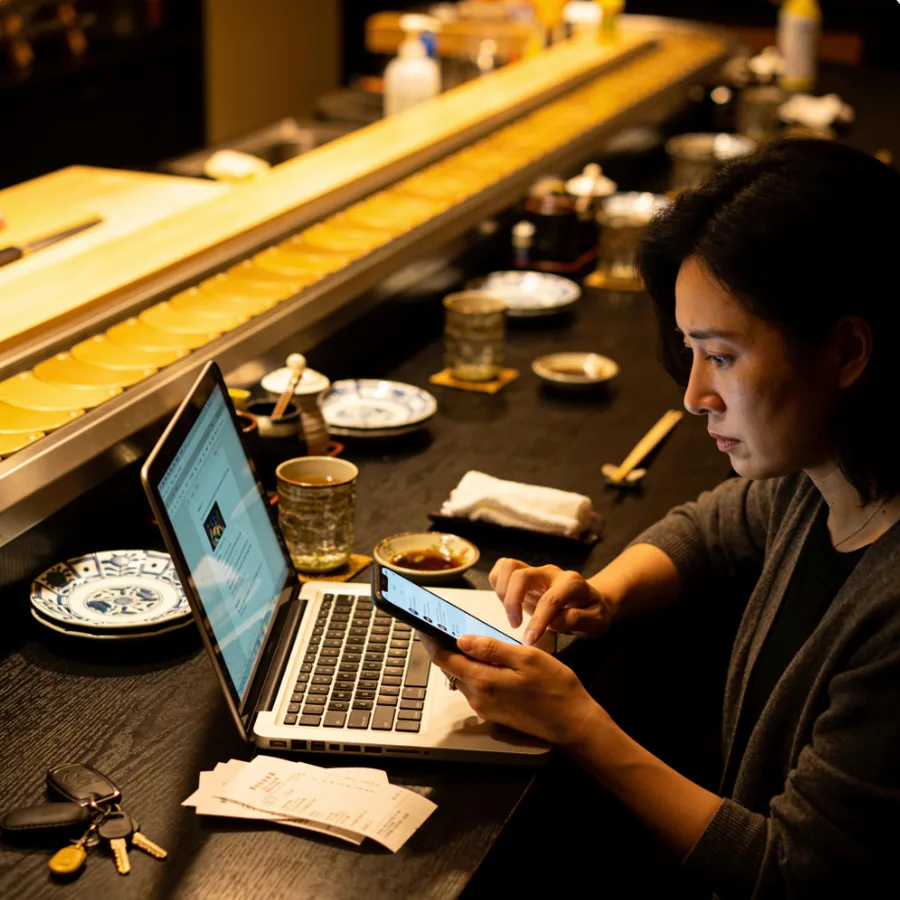 A restaurant owner at her sushi-bar counter late at night, phone in hand and laptop open, handling her own social media